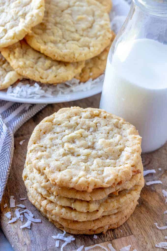 Coconut Cookies stacked on board with milk and plate of cookies in background
