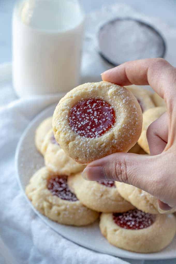 hand holding one thumbprint cookies with plate of cookies and milk in background