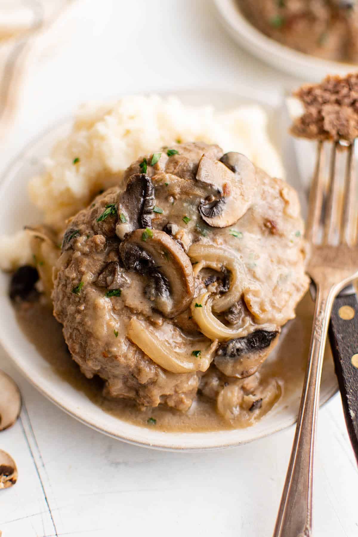 Salisbury steak covered in a sauce of sauteed onions and mushrooms, arranged on a plate next to a serving of mashed potatoes. 