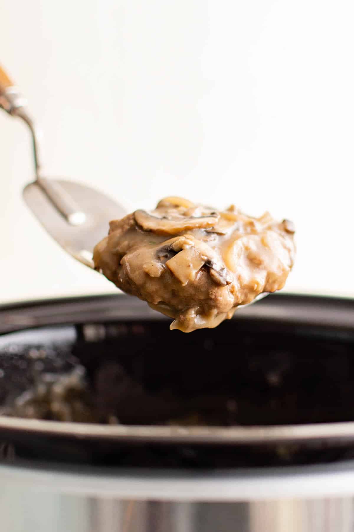 This detailed photograph depicts the fully cooked Salisbury steak patty on a spatula suspended above the crockpot. The sauce glistens in the light with the sliced onions and mushrooms on top of the patty. 