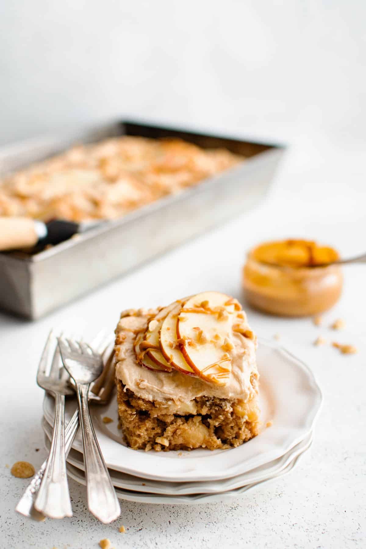 apple peanut butter cake on a plate with forks and toped with peanut butter frosting, sliced apples, and a drizzle of peanut butter