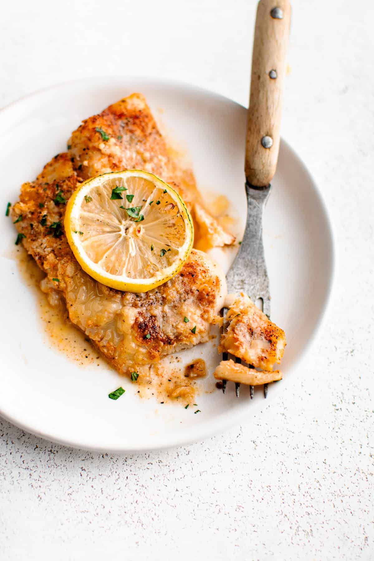 Lemon Garlic Butter Baked Fish on a plate with a fork and garnished with a slice of lemon