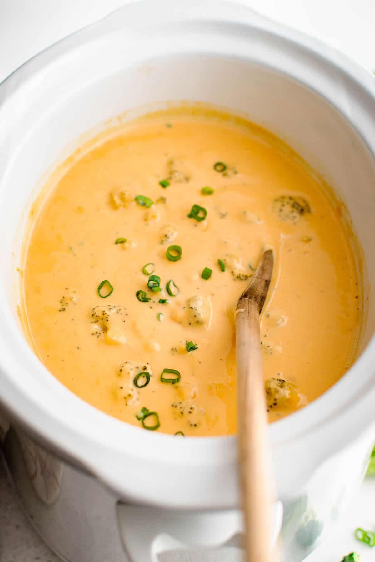 crockpot broccoli cheese sauce in a slow cooker pot with a wooden spoon inserted into it, garnished with green onions