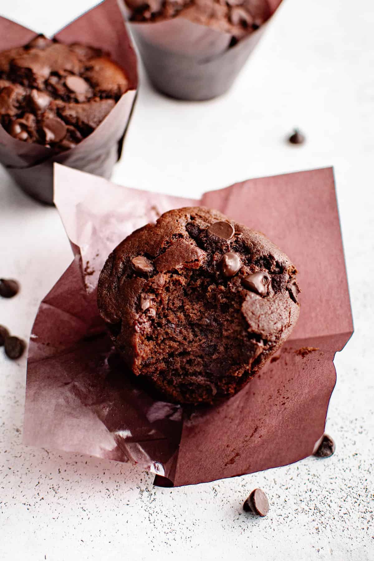 a double chocolate muffin atop its opened wrapper. a bite has been taken out of it revealing the rich chocolately texture inside the muffin