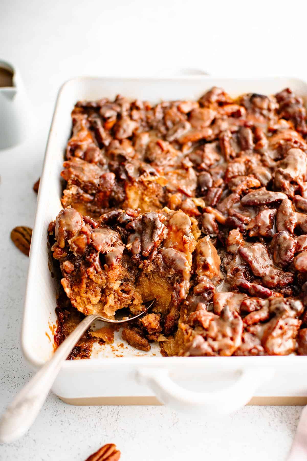 Sticky Bun French Toast Casserole in a baking dish with a large spoon inserted under the casserole. A large portion has been removed so the rich texture of the casserole can be seen